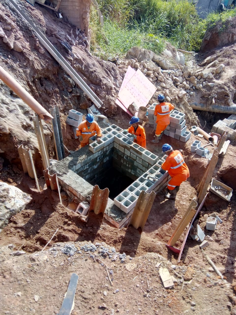 Quatro trabalhadores, de macacão laranja, constroem galeria de captação de água da chuva, na avenida Alcides Perazola